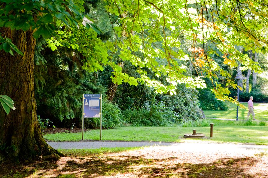 Blick auf die Weiße Quelle im Kurpark in Bad Wildungen. Drumherum sind Bäume und Wiese zu sehen. Vor der Quelle führt ein Weg entlang.
