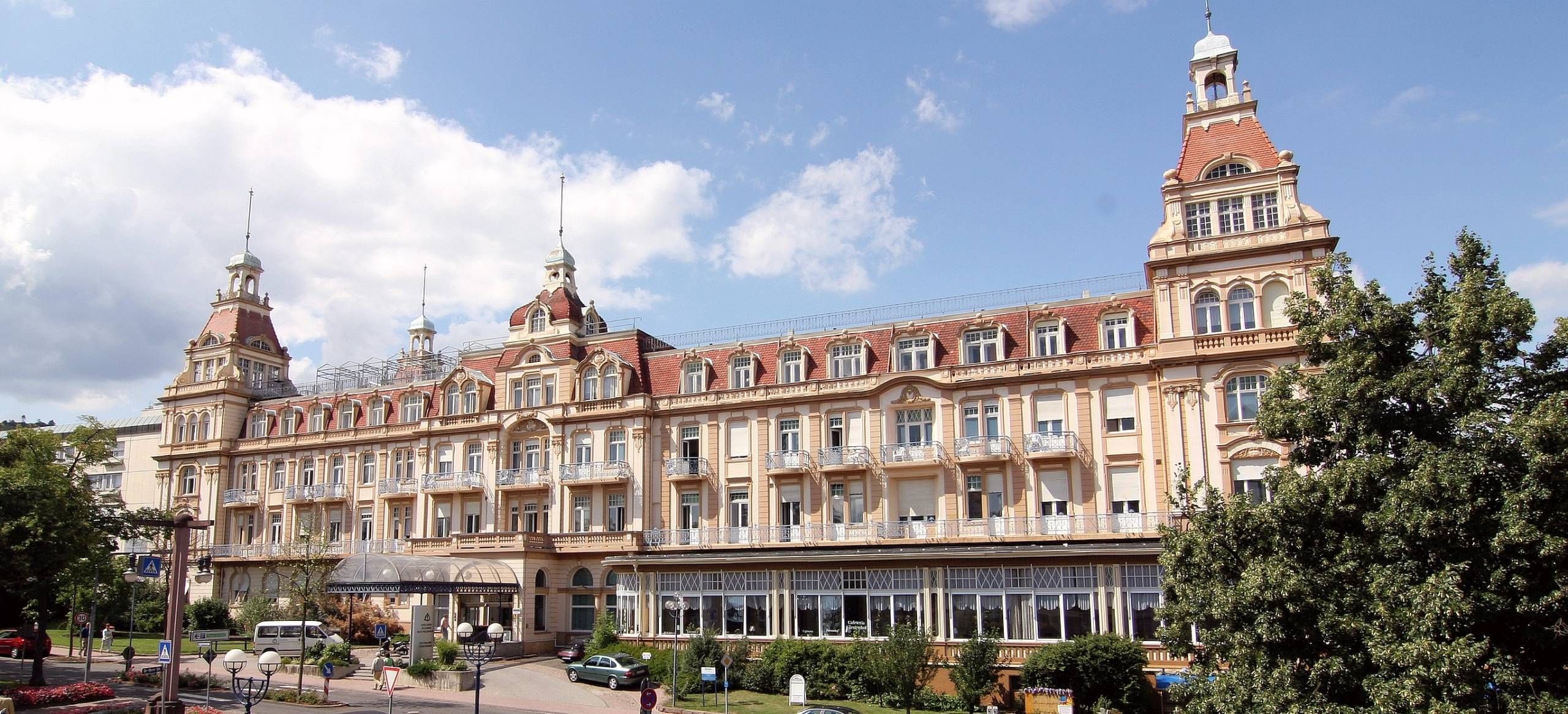 Vorderansicht des Fürstenhofs und der davor verlaufenen Brunnenallee mit Autos und ein paar Fußgängern.