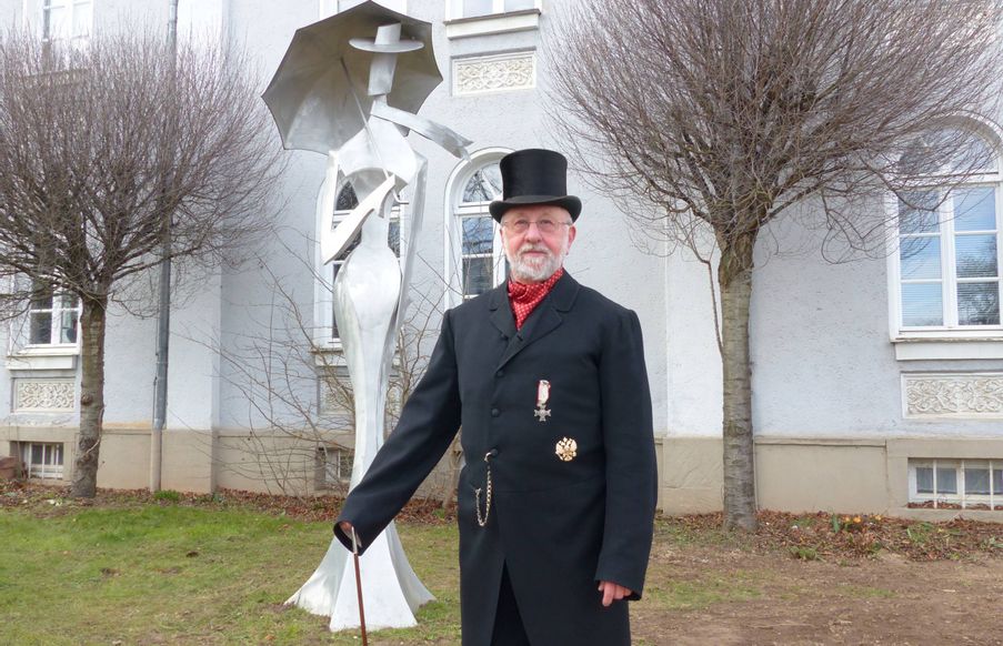 Zu sehen in ein Gästeführer, der vor einen metallenen Statue und einem Gebäude steht, gekleidet im Anzug und mit Zylinder als Graf Korff.