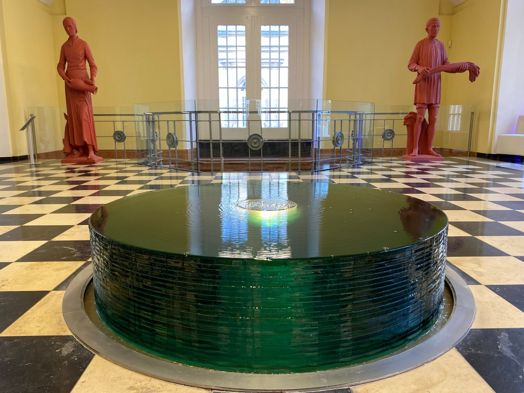 Quellendom in der Wandelhalle Bad Wildungen mit Blick auf einen grünen Brunnen. Im Hintergrund stehen rechts und links zwei rote Statuen und zwischen Ihnen befindet sich ein Geländer.