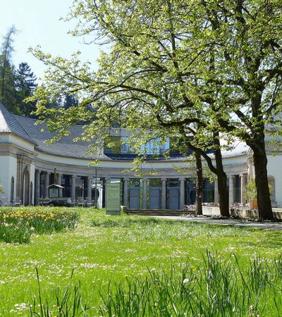 Blick auf die Wandelhalle Bad Wildungen mit grüner Wiese und Bäumen im Vordergrund.
