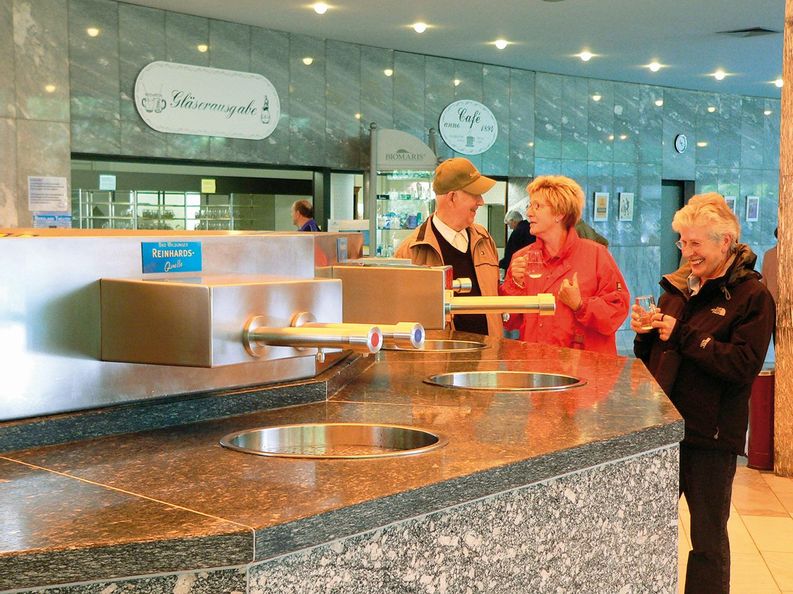 Quellenausschank der Wandelhalle Reinhardshausen. Zwei Frauen und ein Mann stehen hinter dem Ausschank und trinken das Heilwasser. Im Hintergrund des Bildes ist die Gläserausgabe zu sehen.