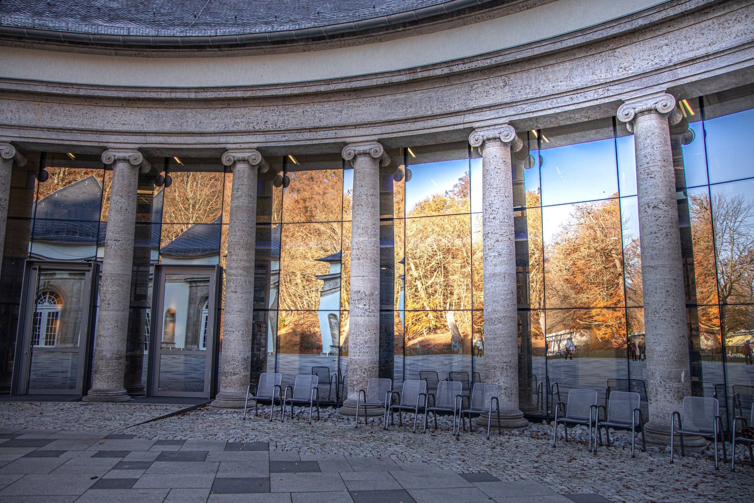 Nahaufnahme auf einen Teil der Vorderseite der Wandelhalle Bad Wildungen bei sonnigem Wetter. In den großen Fenstern, die sich zwischen den großen Säulen befinden, spiegeln sich die herbstlichen Bäume. Vor der Wandelhalle stehen Stühle.