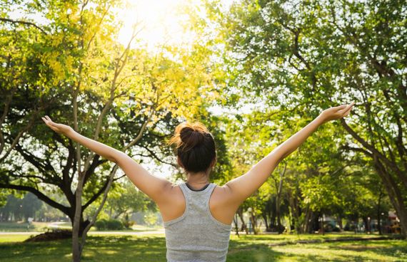 Eine Frau steht mit dem Rücken und ausgestreckten Armen ins Bild auf einer Wiese. Im Hintergrund sind viele grünen Bäume im Sonnenschein zu sehen.