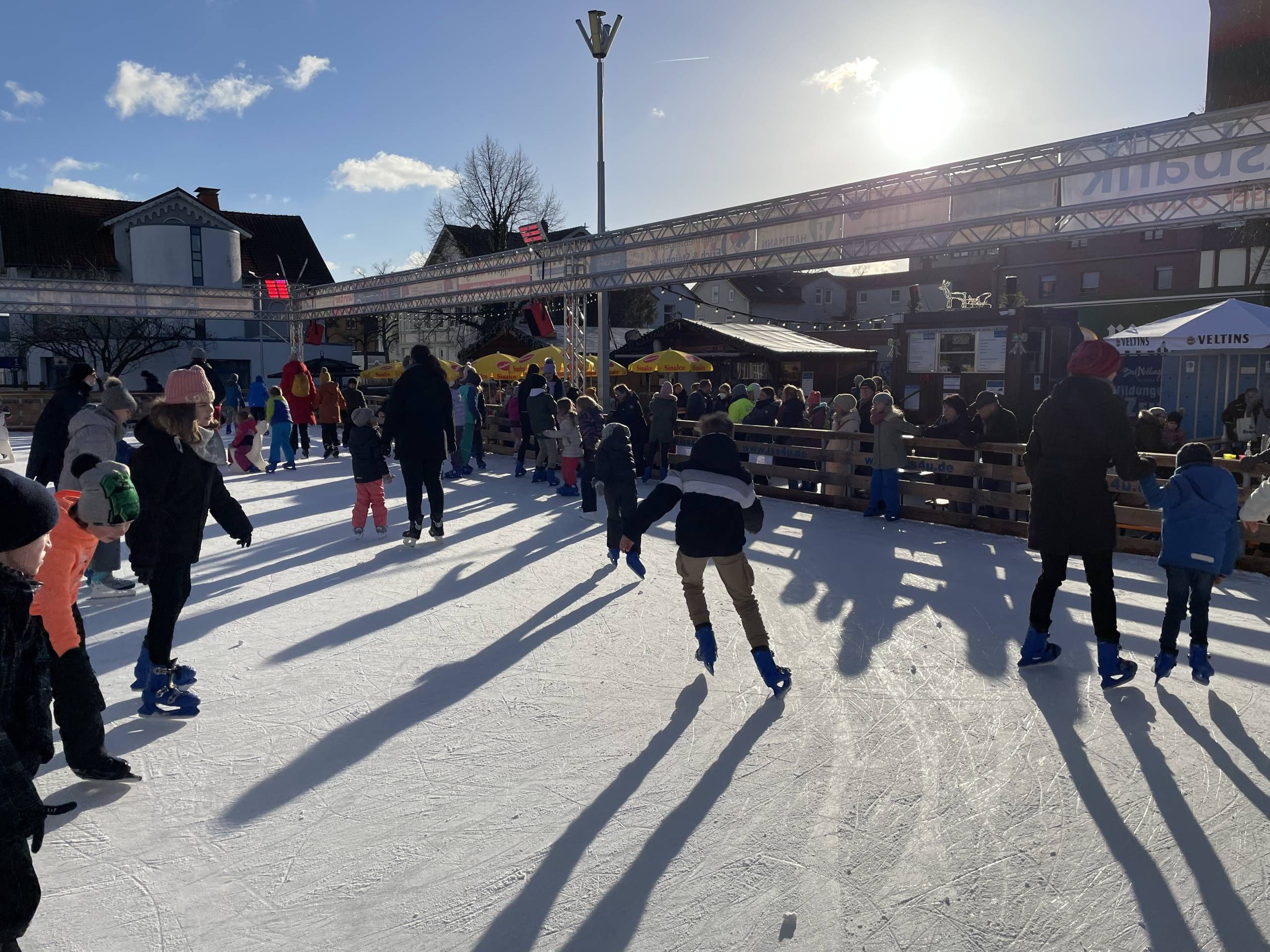 Viele Kinder und ein paar Erwachsene fahren auf der Eisfläche der Bad Wildunger Eis-Zeit mit Schlittschuhen. Die Sonne scheint und im Hintergrund sind ein paar Buden zu sehen.
