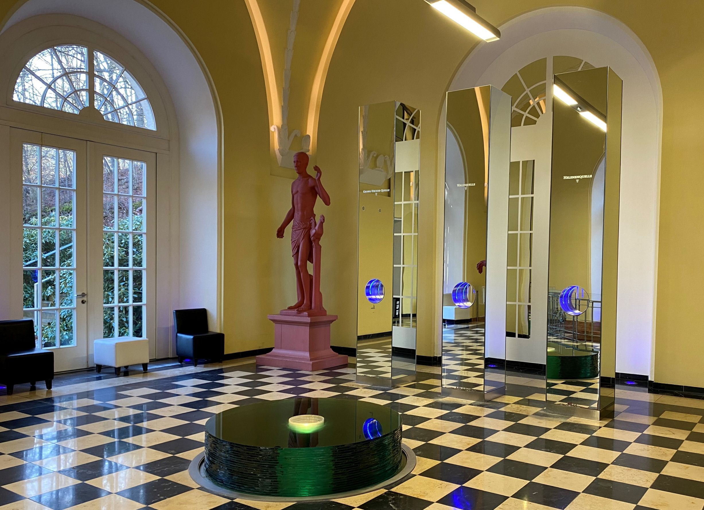 Blick in den Quellendom der Wandelhalle Bad Wildungen. Zu sehen sind die Zapfanlagen des Heilwassers sowie ein rote historische Figur und ein kleiner Quellenbrunnen.