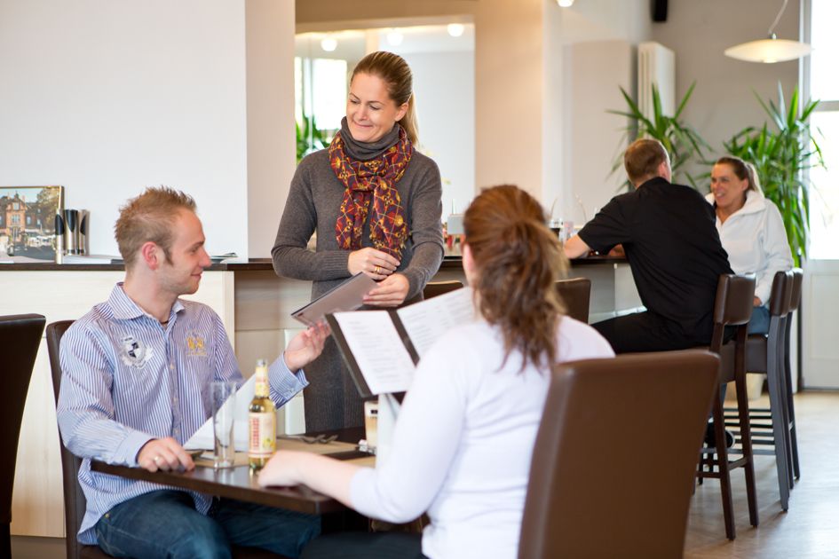 Auf dem Foto sitzen ein Mann und eine Frau an einem Tisch im Restaurant. Vor ihnen steht eine weitere Frau, die dem Mann gerade die Karte reicht. im Hintergrund sitzen ein Mann und eine Frau am Tresen und unterhalten sich.