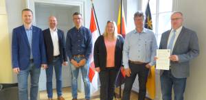 6 Beamte stehen im Rathaus und blicken in die Kamera. Der Bürgermeister hält ein Dokument in die Kamera.