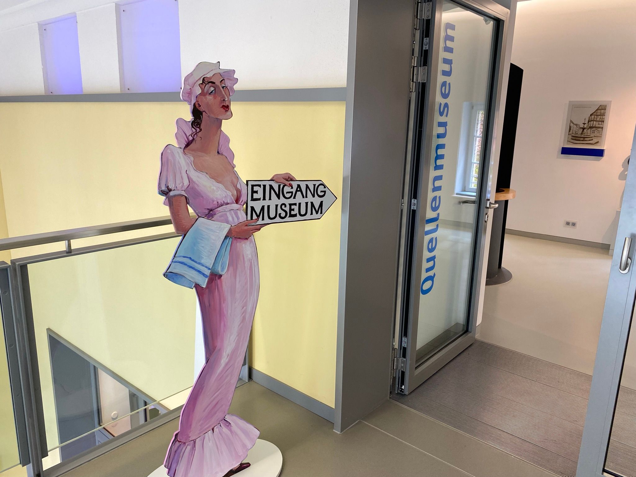 Ein lebensgroßes Schild einer historisch gekleideten Frau, die ein Schild hoch hält, auf dem Eingang Museum steht. Rechts davon ist eine Tür zum Quellenmuseum zu sehen.