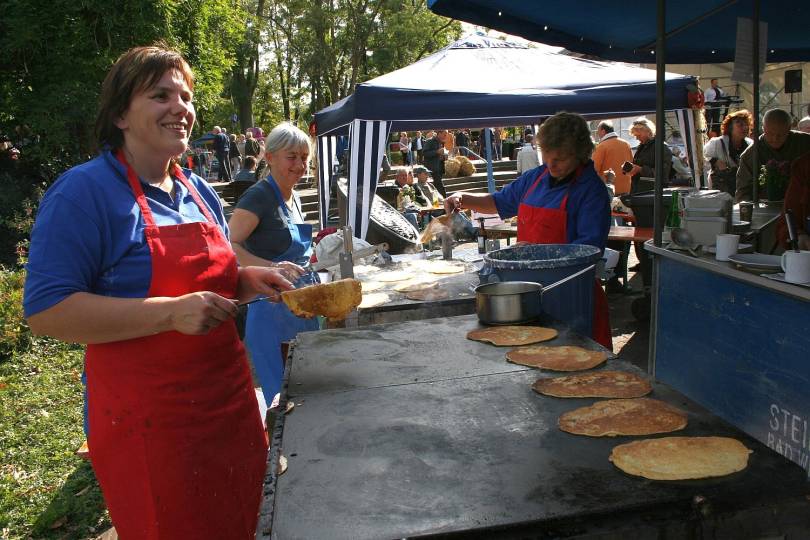 Zu sehen sind drei Personen, die Schepperlinge auf einer großen Grillplatte machen. Im Hintergrund sind viele Menschen auf dem Kartoffelfest im Kurpark zu sehen.