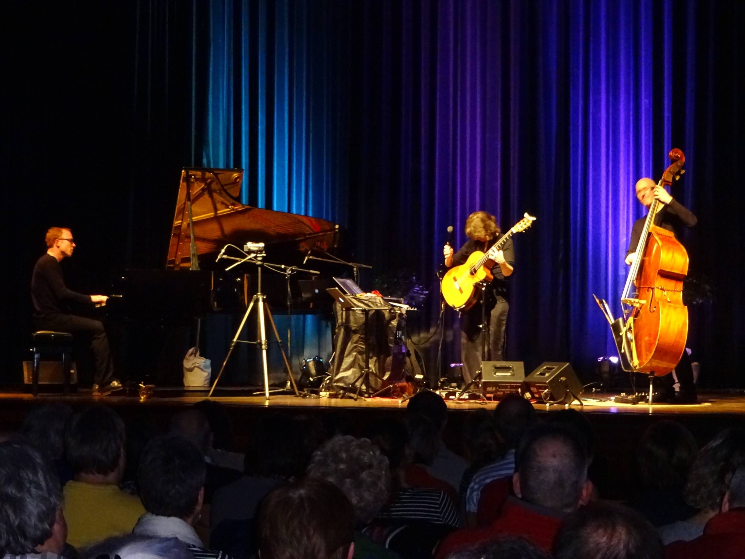 Drei Musiker auf der Bühne des Quellensaals in der Wandelhalle mit verschiedenen Instrumenten, wie ein Klavier. Im Vordergund sind Menschen zu sehen, die vor der Bühne sitzen.