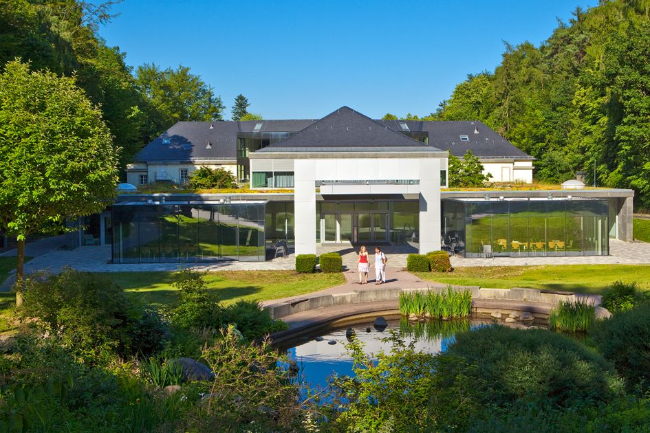 Blick auf die Rückseite der Wandelhalle Bad Wildungen bei Sonnenschein. Rechts und links im Bild sind grüne Bäume zu sehen und im Vordergrund ein Teil des Kurparks mit Teichanlage. Zwei Menschen gehen gerade den Weg entlang.