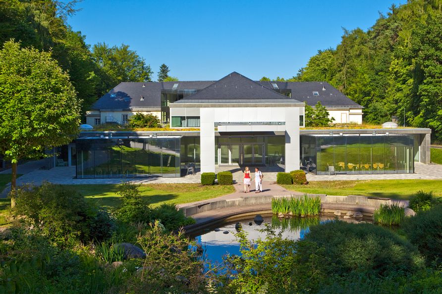 Blick auf die Rückseite der Wandelhalle Bad Wildungen bei Sonnenschein. Rechts und links im Bild sind grüne Bäume zu sehen und im Vordergrund ein Teil des Kurparks mit Teichanlage. Zwei Menschen gehen gerade den Weg entlang.
