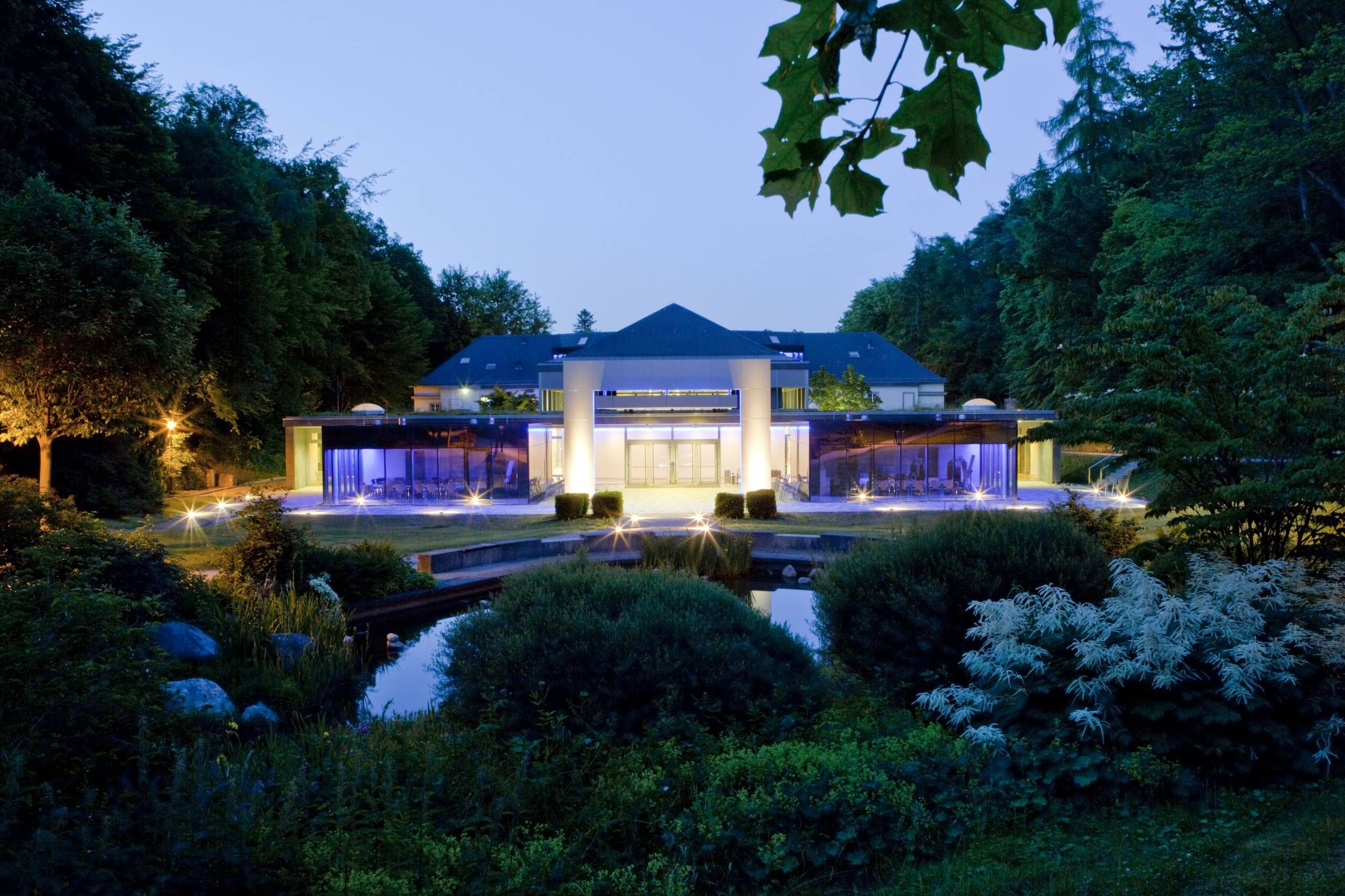 Blick auf die Rückseite der Wandelhalle Bad Wildungen in der Dämmerung. Rechts und links im Bild sind grüne Bäume zu sehen und im Vordergrund ein Teil des Kurparks mit Teichanlage. Die beiden Seminarräume sowie der Eingangsbereich der Wandelhalle sind beleuchtet.
