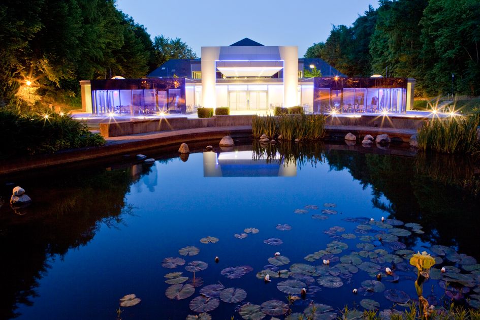 Blick auf die Rückseite der Wandelhalle Bad Wildungen in der Dämmerung. Rechts und links im Bild sind grüne Bäume zu sehen und im Vordergrund ein Teil des Kurparks mit Teichanlage. Die beiden Seminarräume sowie der Eingangsbereich der Wandelhalle sind beleuchtet.