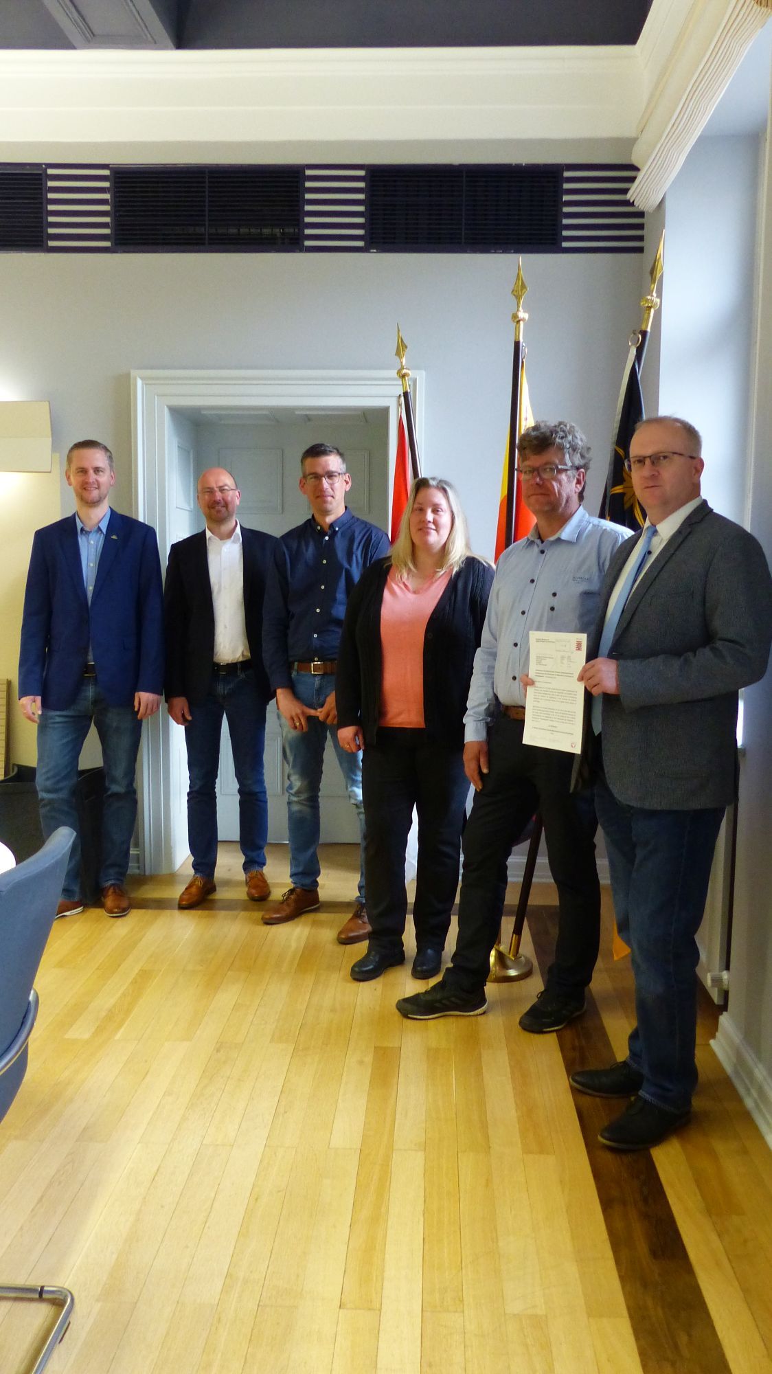 6 Beamte stehen im Rathaus und blicken in die Kamera. Der Bürgermeister hält ein Dokument in die Kamera.