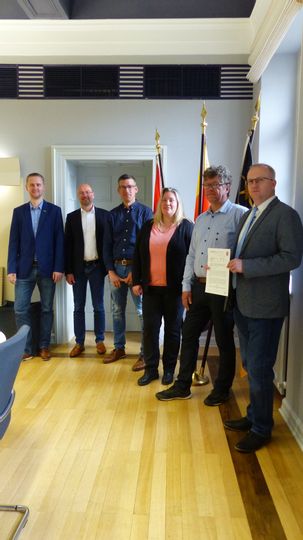 6 Beamte stehen im Rathaus und blicken in die Kamera. Der Bürgermeister hält ein Dokument in die Kamera.