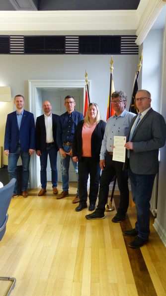 6 Beamte stehen im Rathaus und blicken in die Kamera. Der Bürgermeister hält ein Dokument in die Kamera.