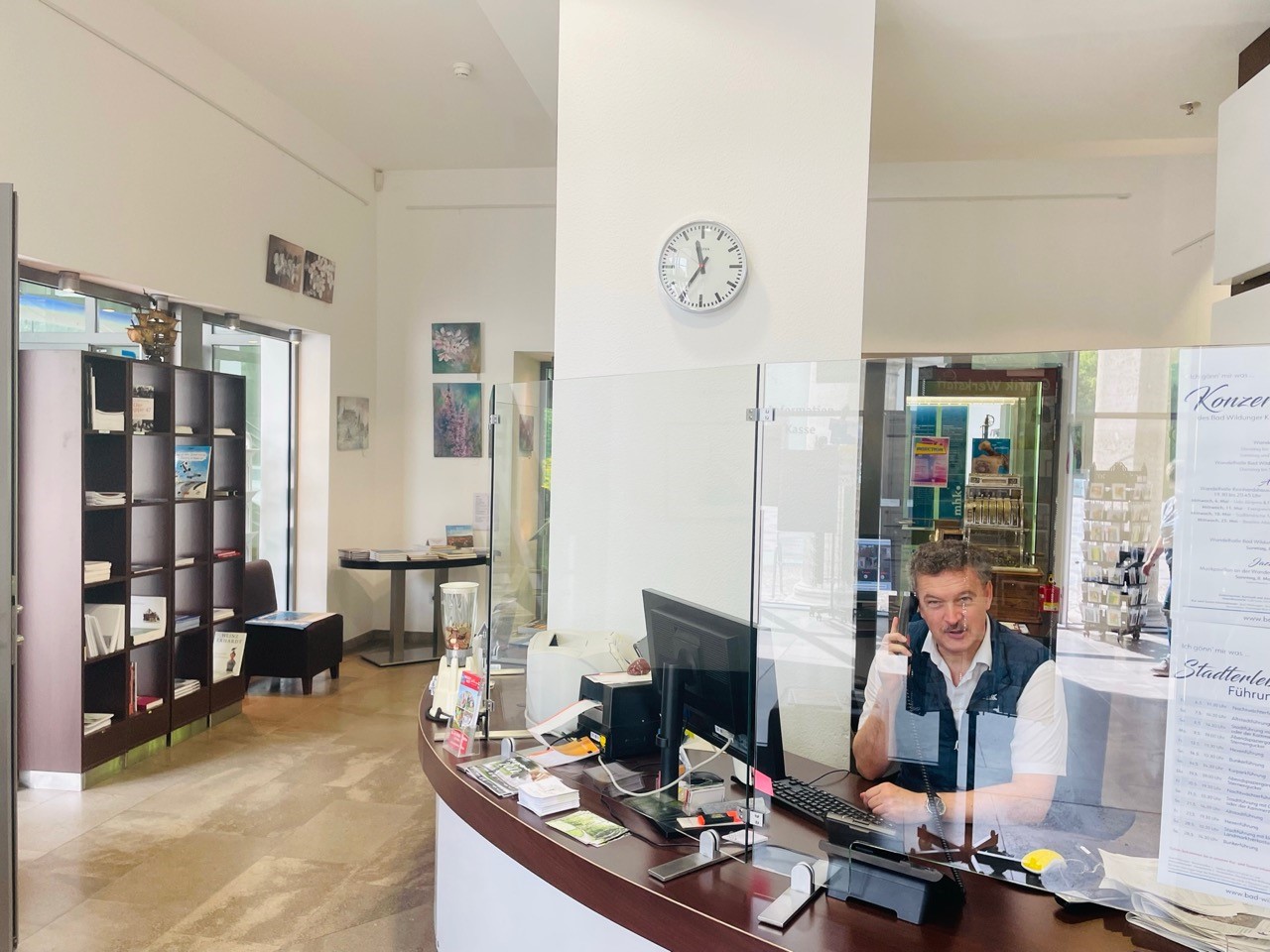 Blick in den Infopoint der Wandelhalle Bad Wildungen. Ein Mitarbeiter sitzt rechts im Bild an der Rezeption und telefoniert gerade. Im linken Teil ist ein Regal und ein Tisch mit Büchern zu sehen sowie ein kleiner Sessel.