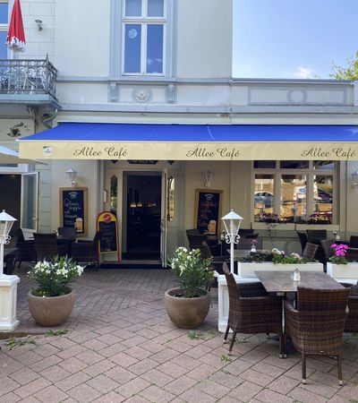 Eingang des Allee Cafés in der Brunnenallee Bad Wildungen. Vor dem Eingang befindet sich ein Terrassenbereich mit Tischen und Stühlen und Blumendekoration.