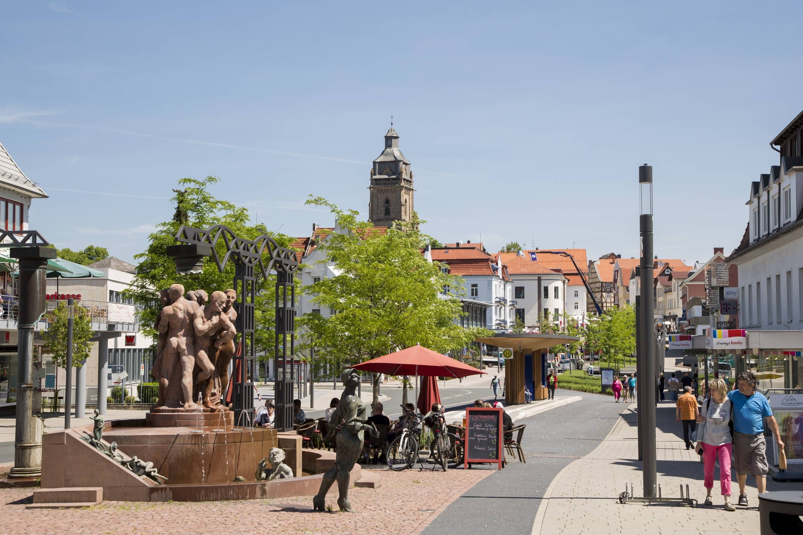Blick auf den Kurschattenbrunnen, die Stadtkirche und die Brunnenstraße mit verschiedenen Einzelhandelsgeschäften. Menschen gehen rechts auf dem Fußweg. Im Vordergrund ist ein Paar zu sehen, das während des Spazierens in Richtung des Brunnens schaut.
