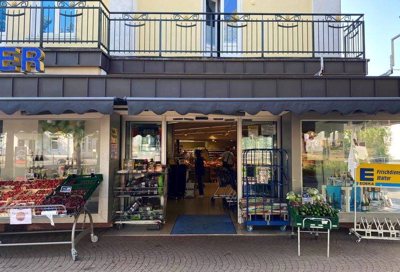 Außenansicht des Eingangs von Edeka Walter. Links vom Eingang sind Obstablagen sowie Körbe mit Grillkohle und Anzünder zu sehen. Rechts im Bild befinden sich eine Gemüseablage mit Gurken sowie Fahrradständer.
