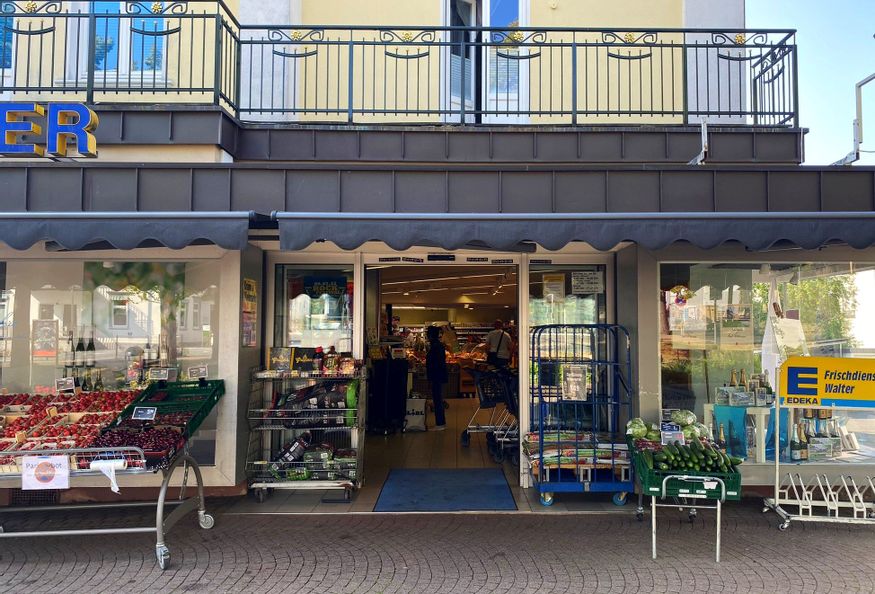 Außenansicht des Eingangs von Edeka Walter. Links vom Eingang sind Obstablagen sowie Körbe mit Grillkohle und Anzünder zu sehen. Rechts im Bild befinden sich eine Gemüseablage mit Gurken sowie Fahrradständer.