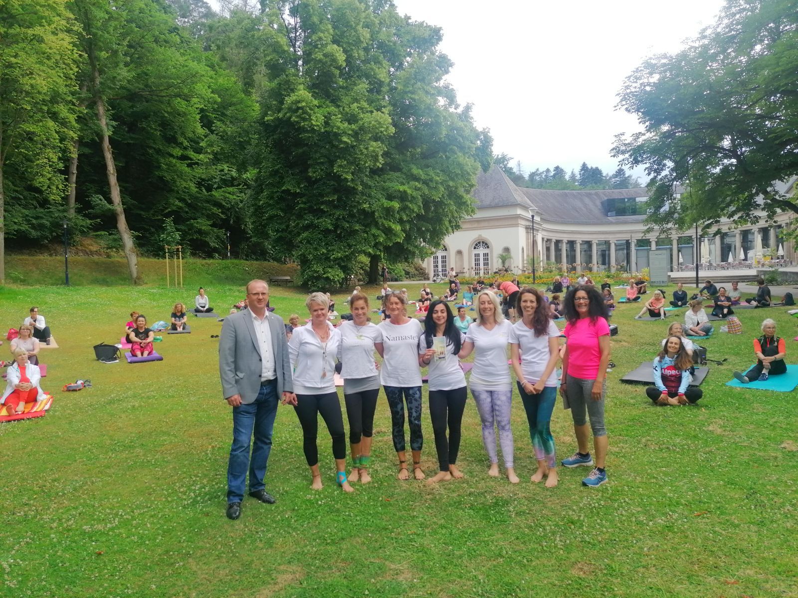 Bild von der Eröffnung des Yogasommers. Im Hintergrund ist die Wandelhalle zu sehen. Viele Teilnehmer sitzen davor auf der Wiese auf ihren Yogamatten. Im Vordergrund sind die sechs Yogalehrerinnen mit dem Bürgermeister Ralf Gutheil und der Stadtmarketing Geschäftsführerin Ute Kühlewind zu sehen.