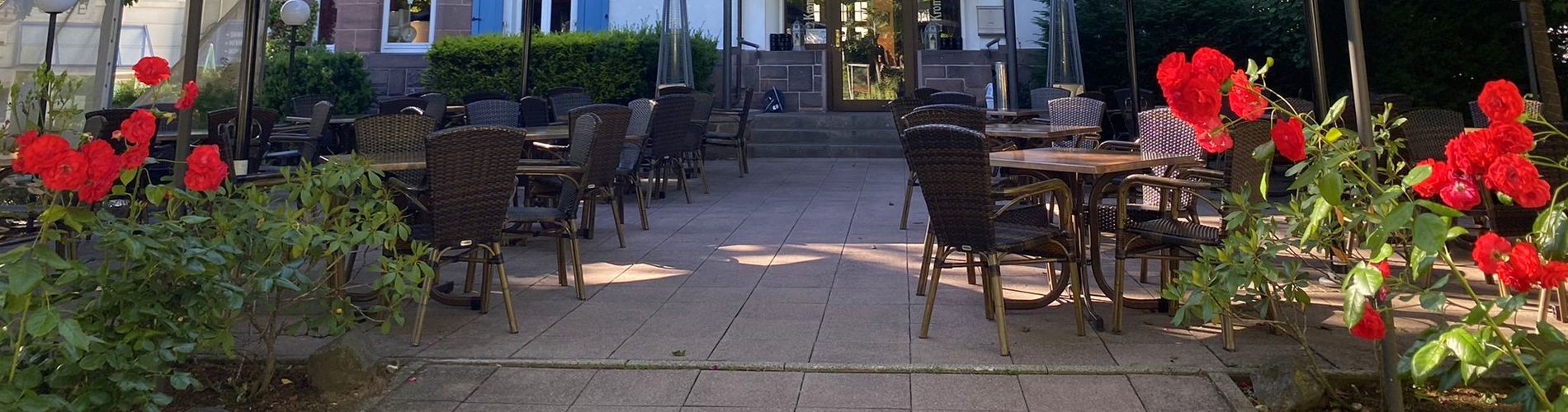 Eine Terrasse mit braunen Tischen und Stühlen und im Vordergrund schöne rote Blumen rechts und links.