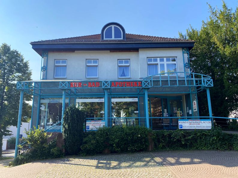 Außenansicht des Gebäudes und Eingangs der Kurbad Apotheke. Im Vordergrund sind leere Parkplätze zu sehen.