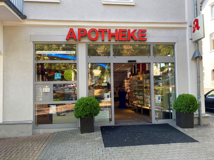 Außenansicht des Eingangs der Löwenapotheke. Neben dem Eingang stehen zwei grüne Sträucher in Töpfen. Die Tür ist geöffnet.