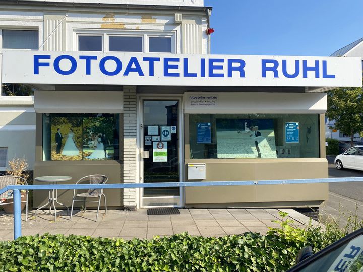 Außenansicht des Eingangs des Fotoateliers Ruhl Links neben dem Eingang stehen zwei Stühle und ein Tisch. Im Schaufenster sind drei Hochzeitsbilder zu sehen.