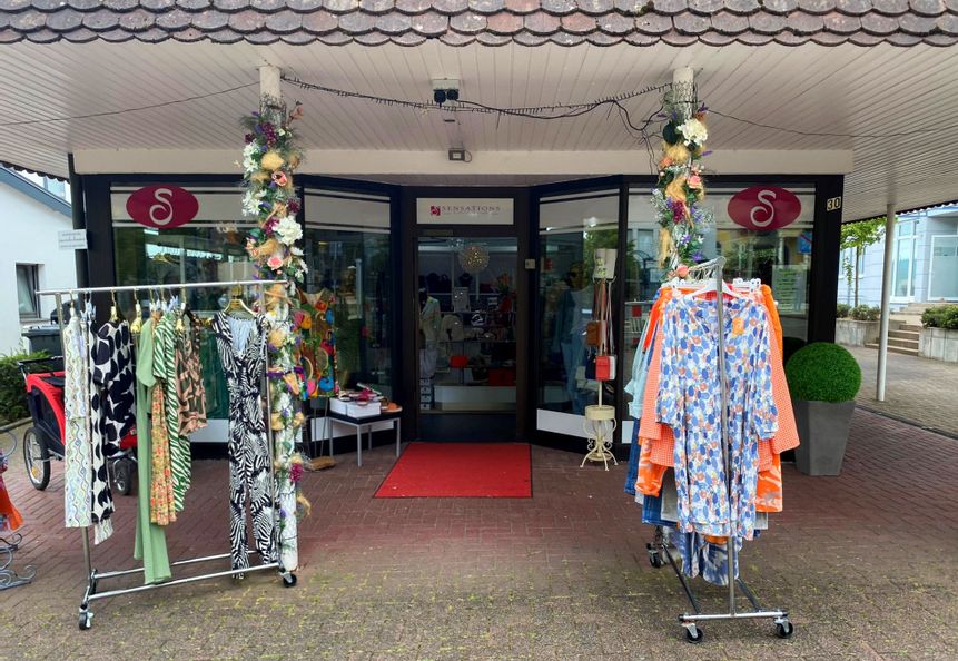 Außenansicht des Bekleidungsgeschäfts sensations. Vor dem Eingang stehen Ständer mit verschiedenen Bekleidungsstücken für Damen.