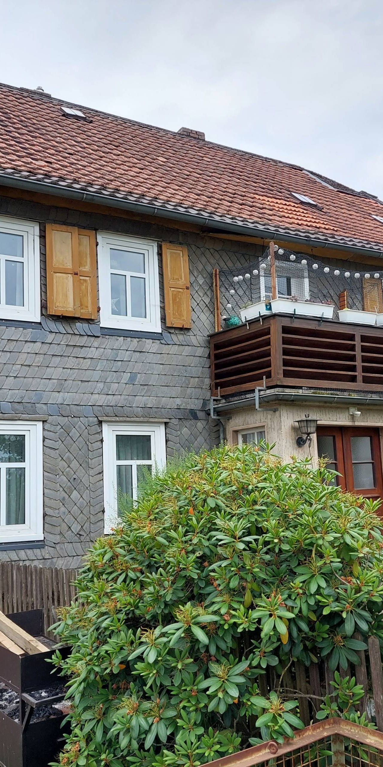 Zu sehen ist das Forsthaus in Hundsdorf. Im Vordergrund ist ein grüner Busch, der über einen Zaun wächst. Ein Balkon ist in der Mitte des Hauses direkt über der Eingangstür.