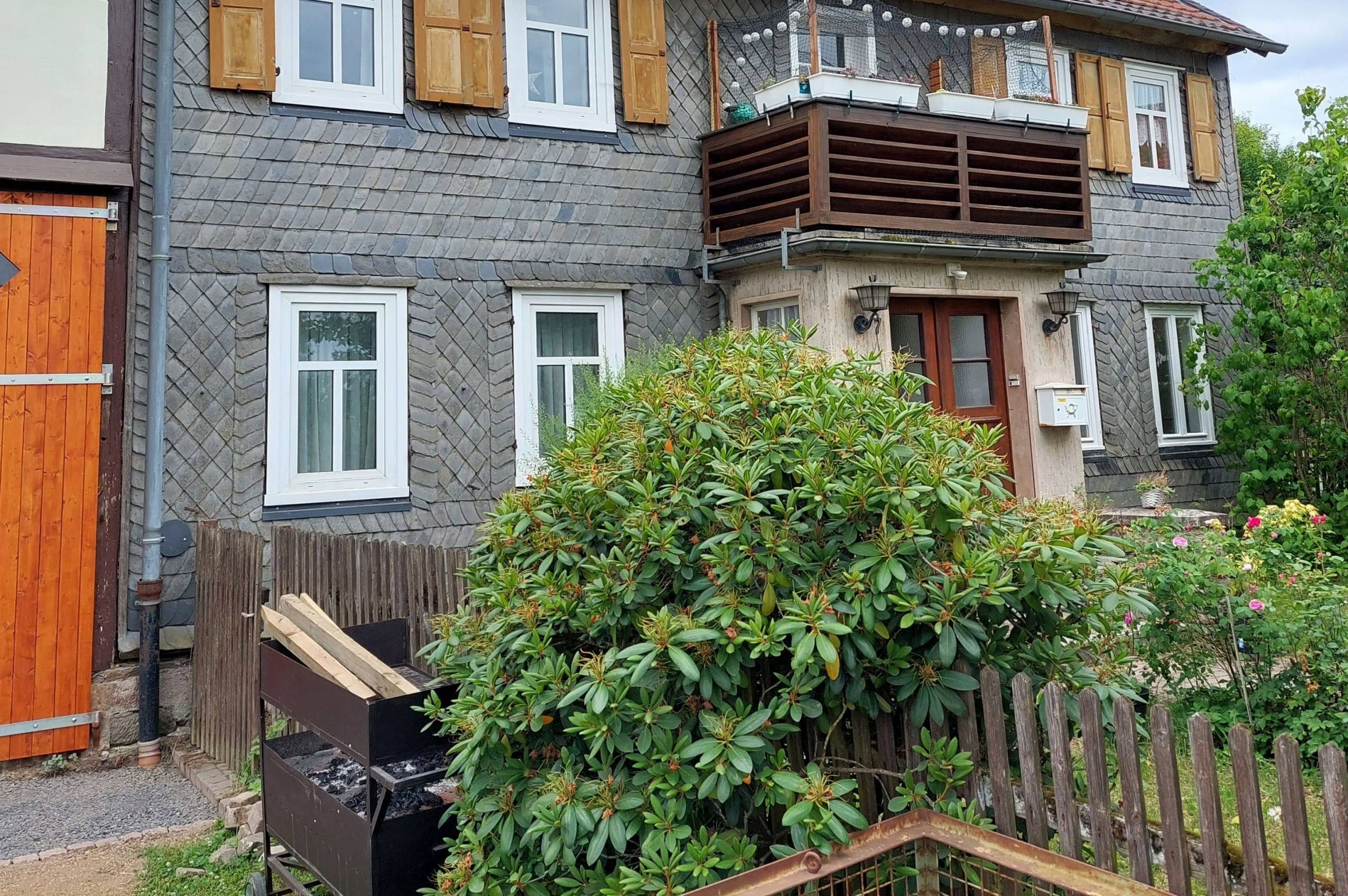 Zu sehen ist das Forsthaus in Hundsdorf. Im Vordergrund ist ein grüner Busch, der über einen Zaun wächst. Ein Balkon ist in der Mitte des Hauses direkt über der Eingangstür.