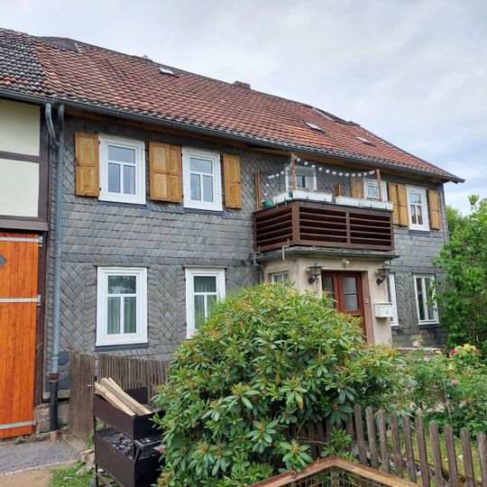 Zu sehen ist das Forsthaus in Hundsdorf. Im Vordergrund ist ein grüner Busch, der über einen Zaun wächst. Ein Balkon ist in der Mitte des Hauses direkt über der Eingangstür.