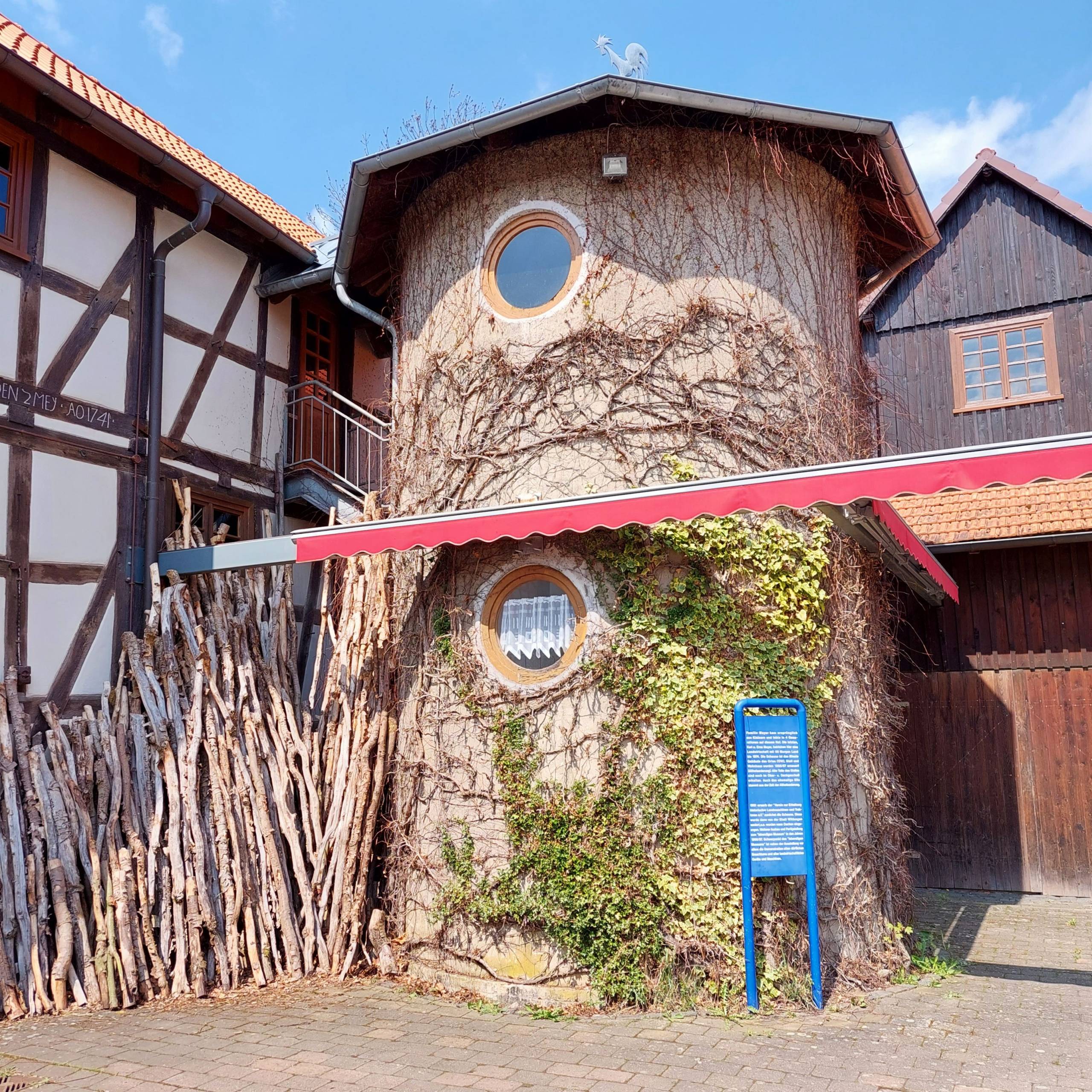 Zu sehen ist das Lebendige Museum in Odershausen. Der mittige Teil des Gebäudes sieht aus wie ein Baumstamm. Links sieht man ein weiß braunes Fachwerkhaus. Auf der rechten Seite des Bildes sieht man ein Haus aus Holz.