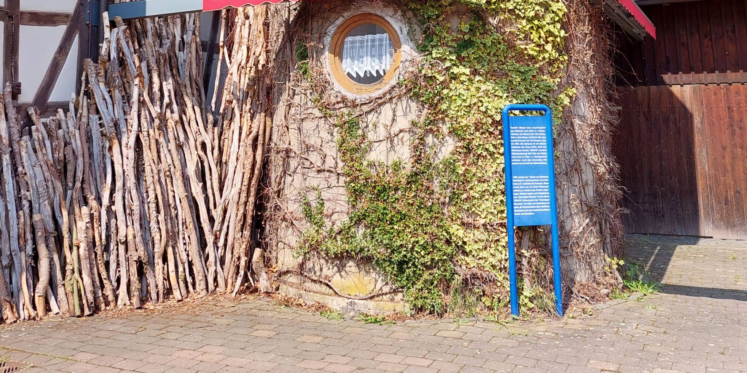 Zu sehen ist das Lebendige Museum in Odershausen. Der mittige Teil des Gebäudes sieht aus wie ein Baumstamm. Links sieht man ein weiß braunes Fachwerkhaus. Auf der rechten Seite des Bildes sieht man ein Haus aus Holz.