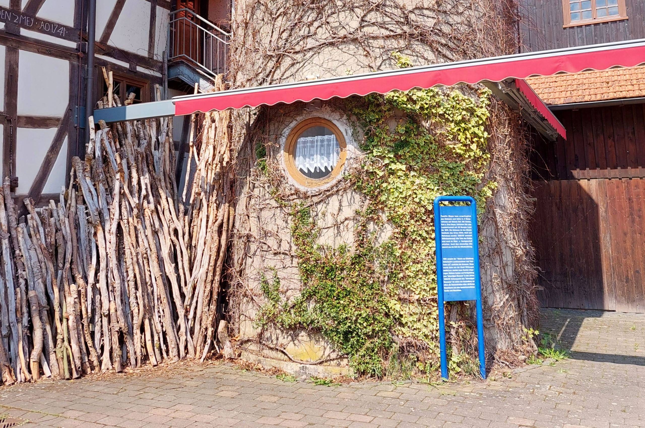 Zu sehen ist das Lebendige Museum in Odershausen. Der mittige Teil des Gebäudes sieht aus wie ein Baumstamm. Links sieht man ein weiß braunes Fachwerkhaus. Auf der rechten Seite des Bildes sieht man ein Haus aus Holz.