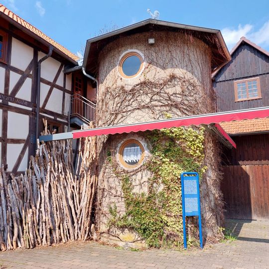 Zu sehen ist das Lebendige Museum in Odershausen. Der mittige Teil des Gebäudes sieht aus wie ein Baumstamm. Links sieht man ein weiß braunes Fachwerkhaus. Auf der rechten Seite des Bildes sieht man ein Haus aus Holz.