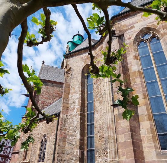 Eine Nahaufnahme vom hinteren Teil, der Stadtkirche in Bad Wildungen. Im Vordergrund ist ein Baum zu sehen, mit grünen Blättern. Im Hintergrund erkennt man die Spitze der Kirche.