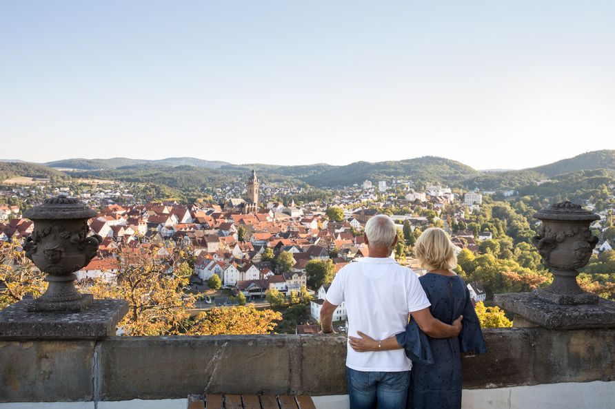 Blick vom Schloss Friedrichstein auf Bad Wildungen bei sonnigem Wetter. In der Mitte ist die Stadtkirche zusehen. Im Vordergrund stehen ein Mann und eine Frau an der Mauer des Schlosses, die sich im Arm halten und auf die Stadt herunterschauen.