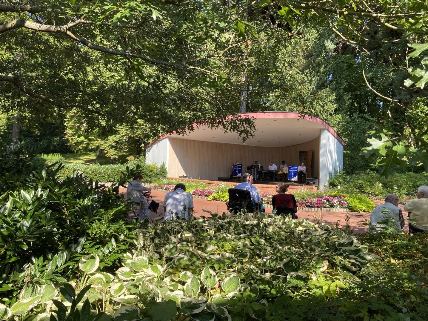 Blick durch die Grünanlage auf den Musikpavillon neben der Wandelhalle Bad Wildungen. Menschen sitzen auf Stühlen davor und hören das Konzert des Kurorchesters.