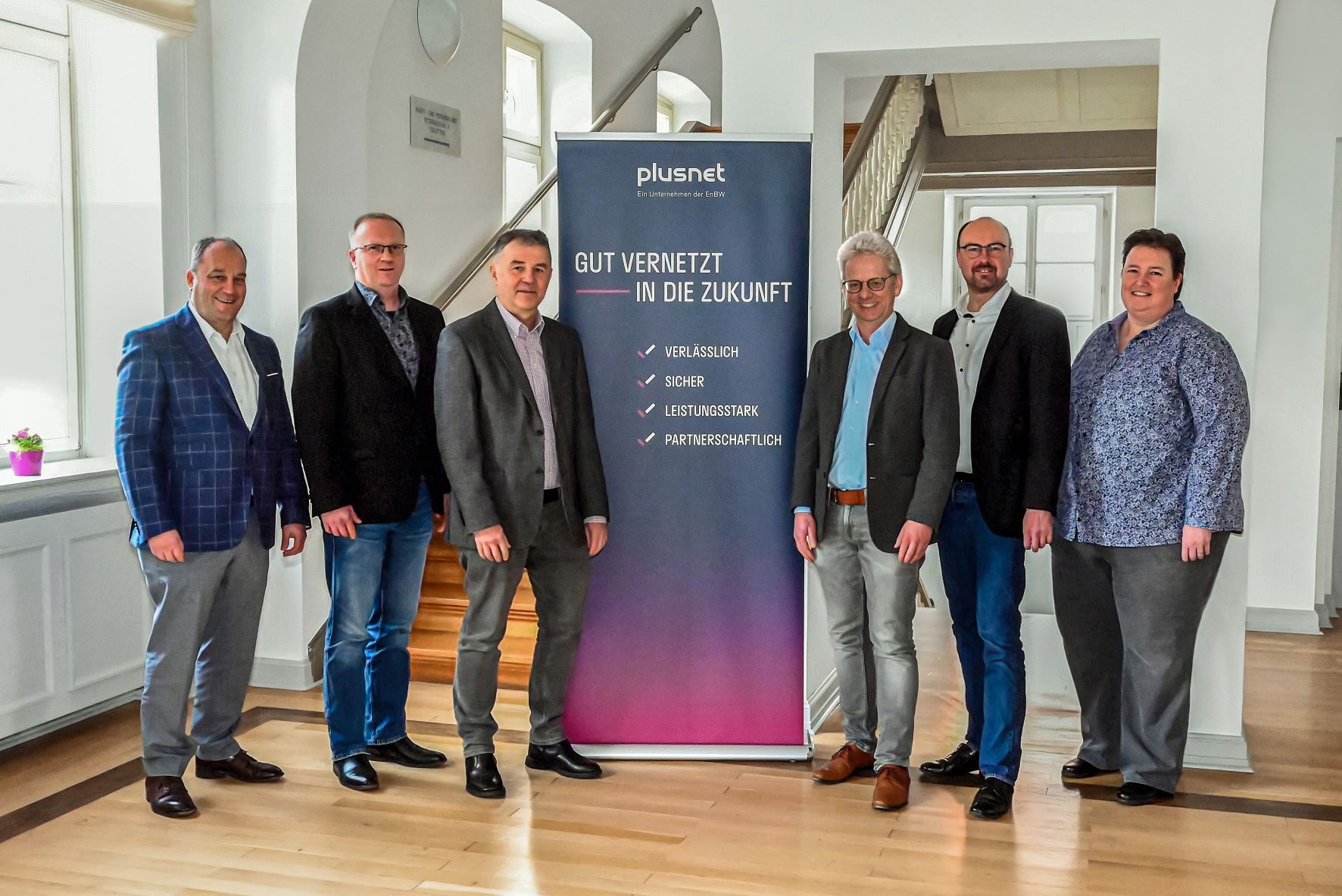 6 Personen stehen im Rathaus neben einem blau-pinken Banner mit der Aufschrift "plusnet Gut vernetzt in die Zukunft".