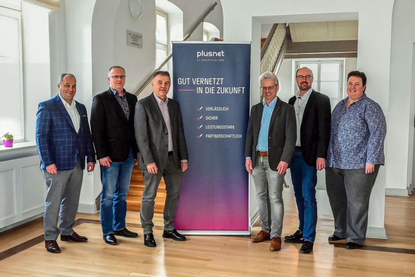 6 Personen stehen im Rathaus neben einem blau-pinken Banner mit der Aufschrift "plusnet Gut vernetzt in die Zukunft".