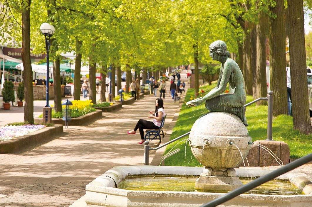 Blick auf die Brunnenallee im Sommer mit der Brunnennymphe im Vordergrund.
