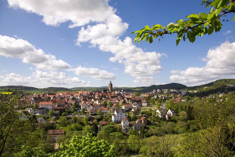 Panoramablick auf Bad Wildungen mit dem Fokus auf die Altstadt und die Stadtkirche. Im Hintergrund ist blauer Himmel mit ein paar Wolken zu sehen. Im Vordergrund sind grüne Bäume.