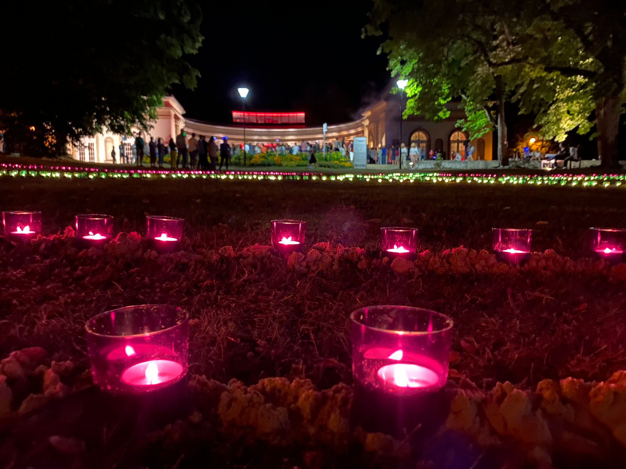 Leuchtende pinke Kerzen auf einer Wiese im Kurpark bei Dunkelheit. Im Hintergrund ist die Wandelhalle mit davorstehenden Menschen zu sehen,