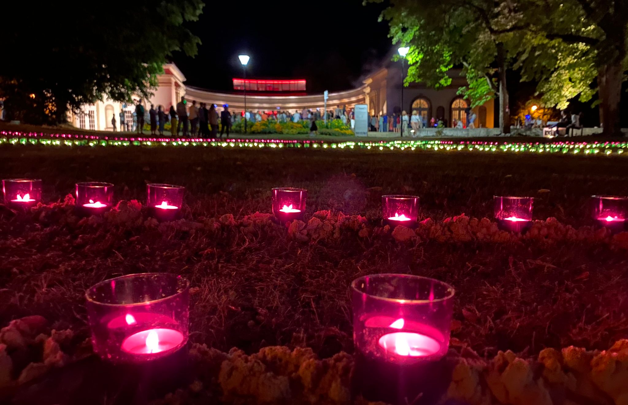 Leuchtende pinke Kerzen auf einer Wiese im Kurpark bei Dunkelheit. Im Hintergrund ist die Wandelhalle mit davorstehenden Menschen zu sehen,