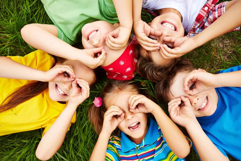 Fünf bunt gekleidete Kinder, die auf der grünen Wiese liegen und sich die Hände wie Brillen um die Augen legen.